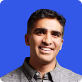 Smiling man with short dark hair wearing a gray button-up shirt against a blue background.